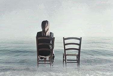 woman with back to camera sitting in wood chair with empty chair next to her in foot of water