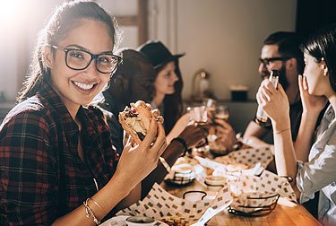 woman in glasses at table with people holding sandwich, looking at camera